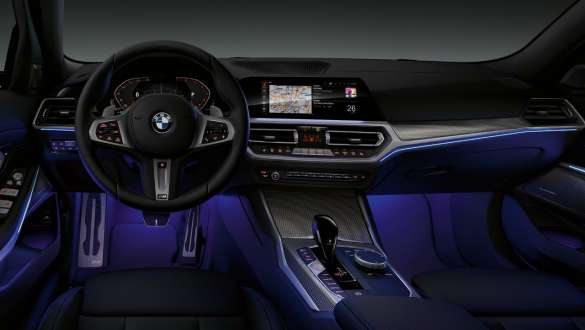 BMW Serie 3 Berlina (G20), Ambient Light. Ripresa anteriore del cockpit dal lato guidatore della BMW Serie 3 Berlina (G20) con vano piedi illuminato di blu grazie all’Ambient Light.