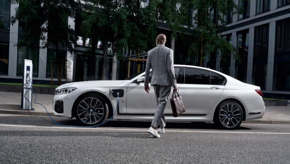 BMW 745e Berlina ibrida Plug-inG11 G12 Mineral White metallizzato vista laterale vicino a colonnina di ricarica BMW 745e Berlina ibrida Plug-inG11 G12 Mineral White metallizzato vista laterale vicino alla colonnina di ricarica con modella