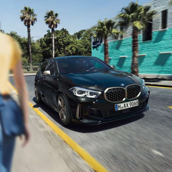 BMW M135i xDrive, vista anteriore a tre quarti. BMW M135i xDrive in Sapphire Black metallizzato, vista anteriore a tre quarti con donna in primo piano.