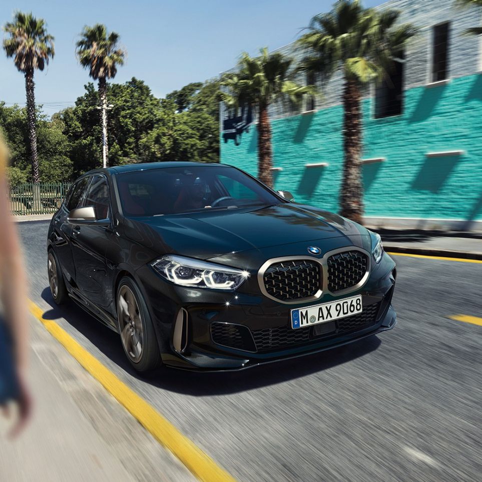 BMW M135i xDrive, vista anteriore a tre quarti F40. BMW M135i xDrive in Sapphire Black metallizzato, vista anteriore a tre quarti con donna in primo piano F40.