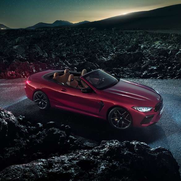 BMW M8 M Competition, vista laterale di tre quarti F91. BMW M8 Competition Cabrio in Motegi Red metallizzato con capote aperta, vista laterale di tre quarti, in sosta di notte sull'asfalto F91.