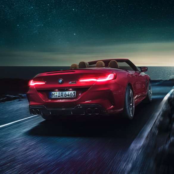 BMW M8 M Competition Cabrio, vista posteriore di tre quarti F91. BMW M8 Competition Cabrio in Motegi Red metallizzato con capote aperta, vista posteriore di tre quarti, in marcia di notte su strada costiera F91.