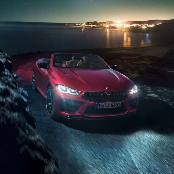 BMW M8 M Competition Cabrio, vista laterale di tre quarti F91. BMW M8 Competition Cabrio in Motegi Red metallizzato con capote aperta, vista frontale di tre quarti, in marcia di notte su strada costiera F91.