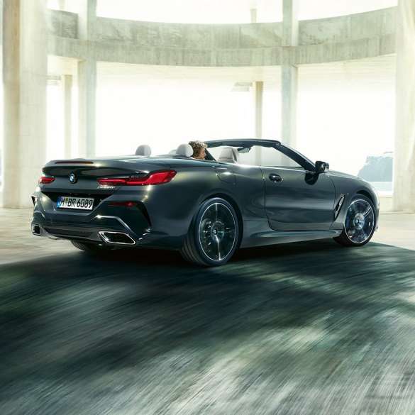 BMW M850i xDrive Cabrio, vista posteriore di tre quarti G14. BMW M850i xDrive Cabrio in Dravit Grey metallizzato con capote aperta, vista posteriore di tre quarti, in marcia attraverso un edificio in calcestruzzo G14.