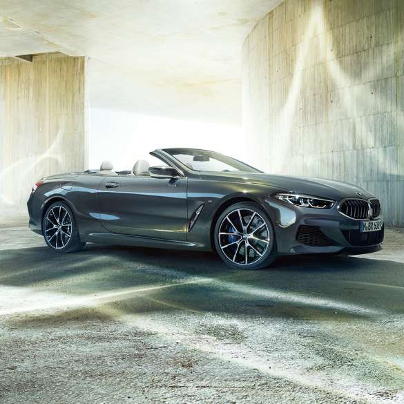 BMW M850i xDrive Cabrio, vista laterale di tre quarti G14. BMW M850i xDrive Cabrio in Dravit Grey metallizzato con capote aperta, vista laterale di tre quarti, in sosta davanti a un edificio in calcestruzzo G14.