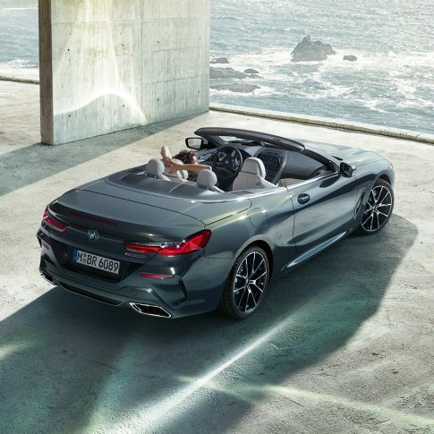 BMW M850i xDrive Cabrio, prospettiva a volo d’uccello G14. BMW M850i xDrive Cabrio in Dravit Grey metallizzato con capote aperta, prospettiva a volo d’uccello, parcheggiata in edificio di calcestruzzo davanti all’oceano G14.