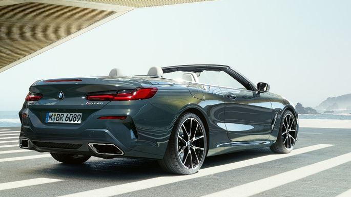 BMW M850i xDrive Cabrio, vista posteriore di tre quarti G14. BMW M850i xDrive Cabrio in Dravit Grey metallizzato con capote aperta, vista posteriore di tre quarti, parcheggiata con sguardo sull’oceano G14.