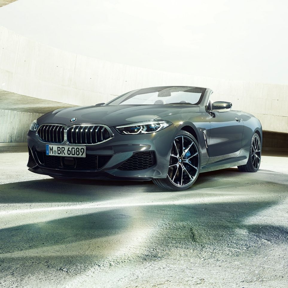 BMW M850i xDrive Cabrio, vista frontale di tre quarti G14. BMW M850i xDrive Cabrio in Dravit Grey metallizzato con capote aperta, vista frontale di tre quarti, in sosta in un edificio in calcestruzzo G14.