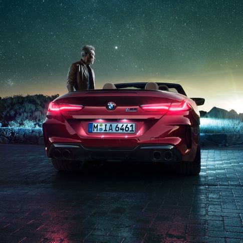 BMW M8 Competition Cabrio, vista posteriore F91. BMW M8 Competition Cabrio in Motegi Red metallizzato con capote aperta, vista posteriore, in sosta con uomo sotto il cielo stellato F91.