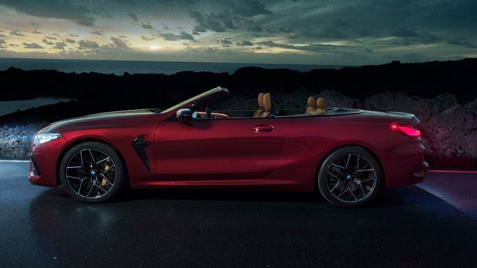 BMW M8 Competition Cabrio, vista laterale F91. BMW M8 Competition Cabrio in Motegi Red metallizzato con capote aperta, vista laterale, in sosta di notte sotto il cielo stellato F91.