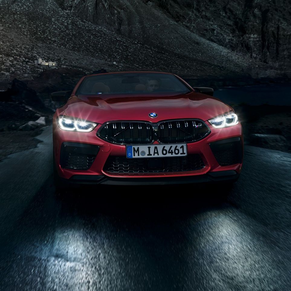 Nuova BMW M8 Competition Cabrio, vista frontale F91. Nuova BMW M8 Competition Cabrio in Motegi Red metallizzato con fari accesi, vista frontale, in marcia di notte F91.