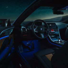 Interni BMW M8 Competition Coupé F92. BMW M8 Competition Coupé, interni di notte con volante in pelle M e luce ambiente blu con porta del guidatore aperta F92.