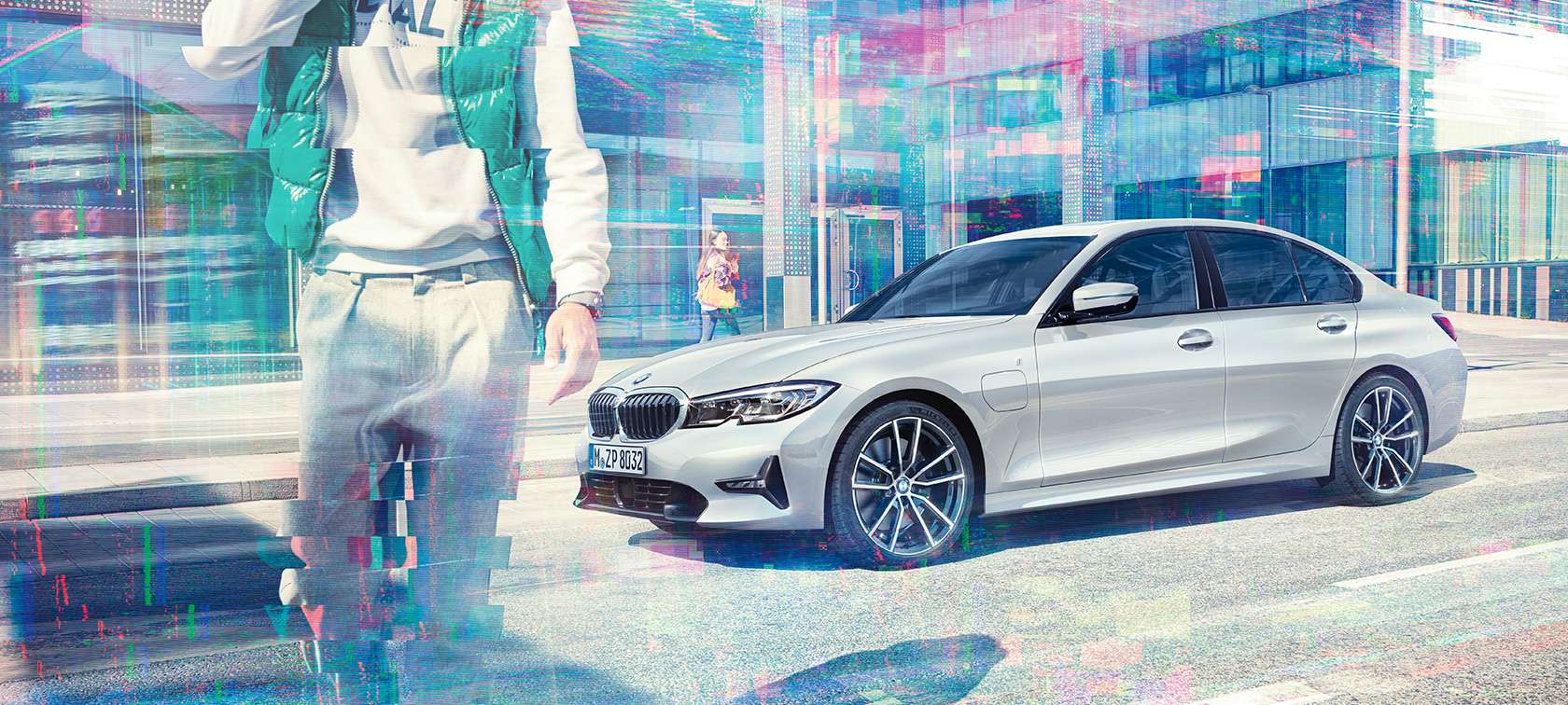 Three-quarter side shot of the standing BMW 330e iPerformance Sedan in front of an urban glass building with walking passengers. BMW 3 Series Sedan, side shot BMW 330e iPerformance.