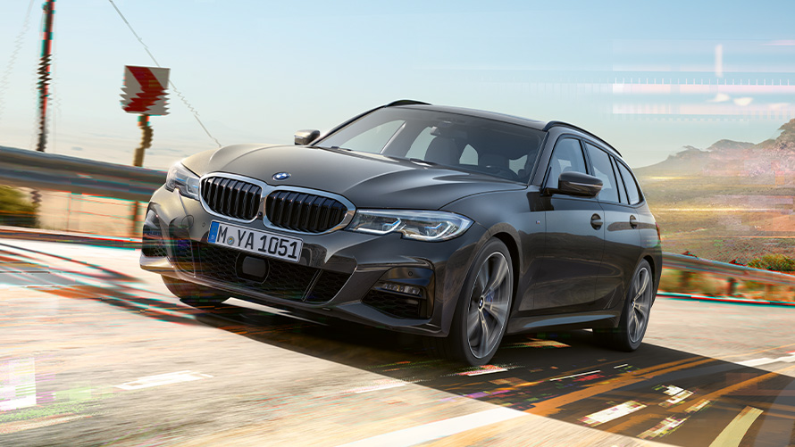 BMW Serie 3 Touring (G21) Dravit Grey, vista anteriore a tre quarti BMW Serie 3 Touring (G21) Dravit Grey, vista anteriore a tre quarti in movimento con sfondo rurale