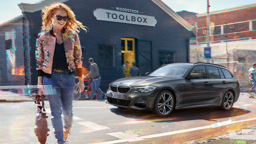 BMW Serie 3 Touring (G21), vista anteriore a tre quarti su strada con donna e ferramenta sullo sfondo Donna attraversa la strada davanti a una BMW Serie 3 Touring (G21) con sullo sfondo una ferramenta.