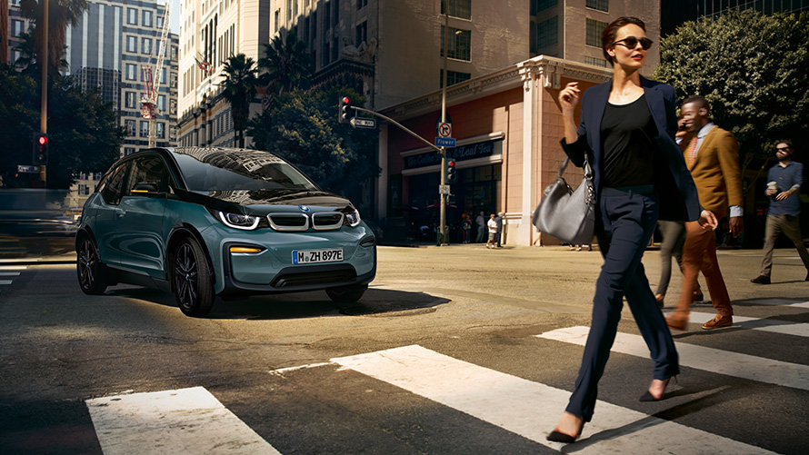 BMW i3 I01 vista anteriore a tre quarti con donna che attraversa sulle strisce pedonali BMW i3 I01 Blue Ridge Mountain metallizzato con profilo in Frozen Grey metallizzato vista frontale a tre quarti con donna che attraversa sulle strisce pedonali