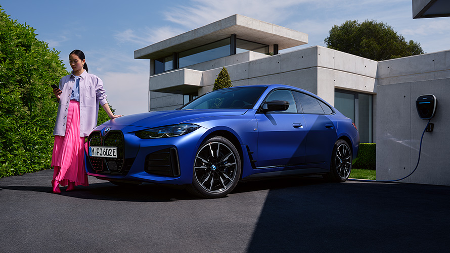 BMW i4 M50 G26 2021 BMW Individual Frozen Portimao Blue metallizzato Vista anteriore a tre quarti ferma con donna davanti a un immobile