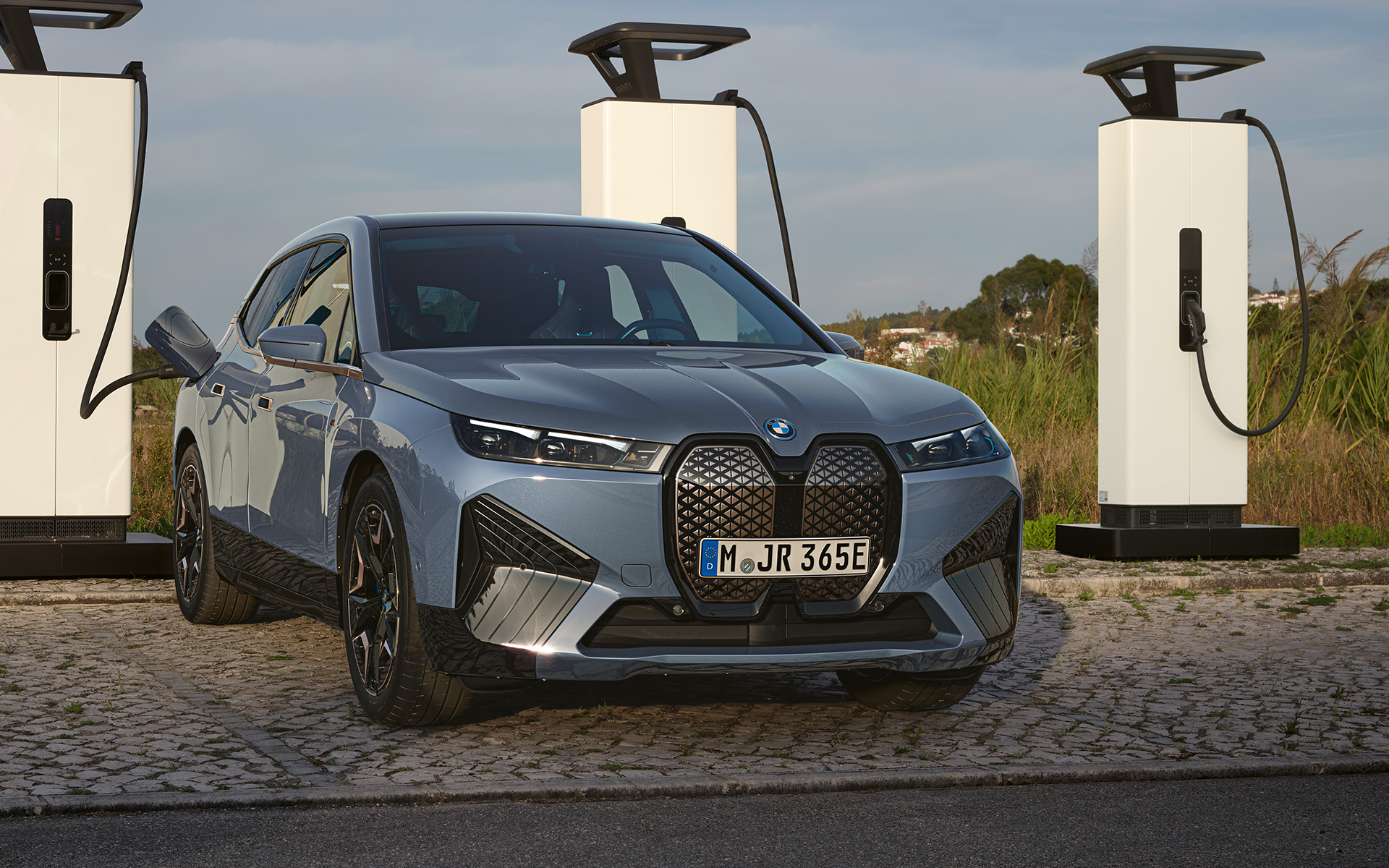 BMW iX M60 i20 2022 SUV elettrico BMW Individual Storm Bay metallizzato Vista anteriore presso una stazione di ricarica High Power