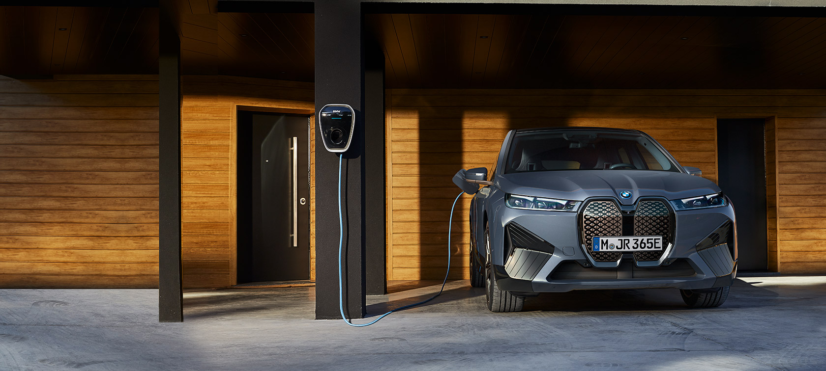 BMW iX M60 i20 2022 SUV elettrico BMW Individual Storm Bay metallizzato Vista anteriore in ricarica davanti ad una casa vicino a una BMW Wallbox