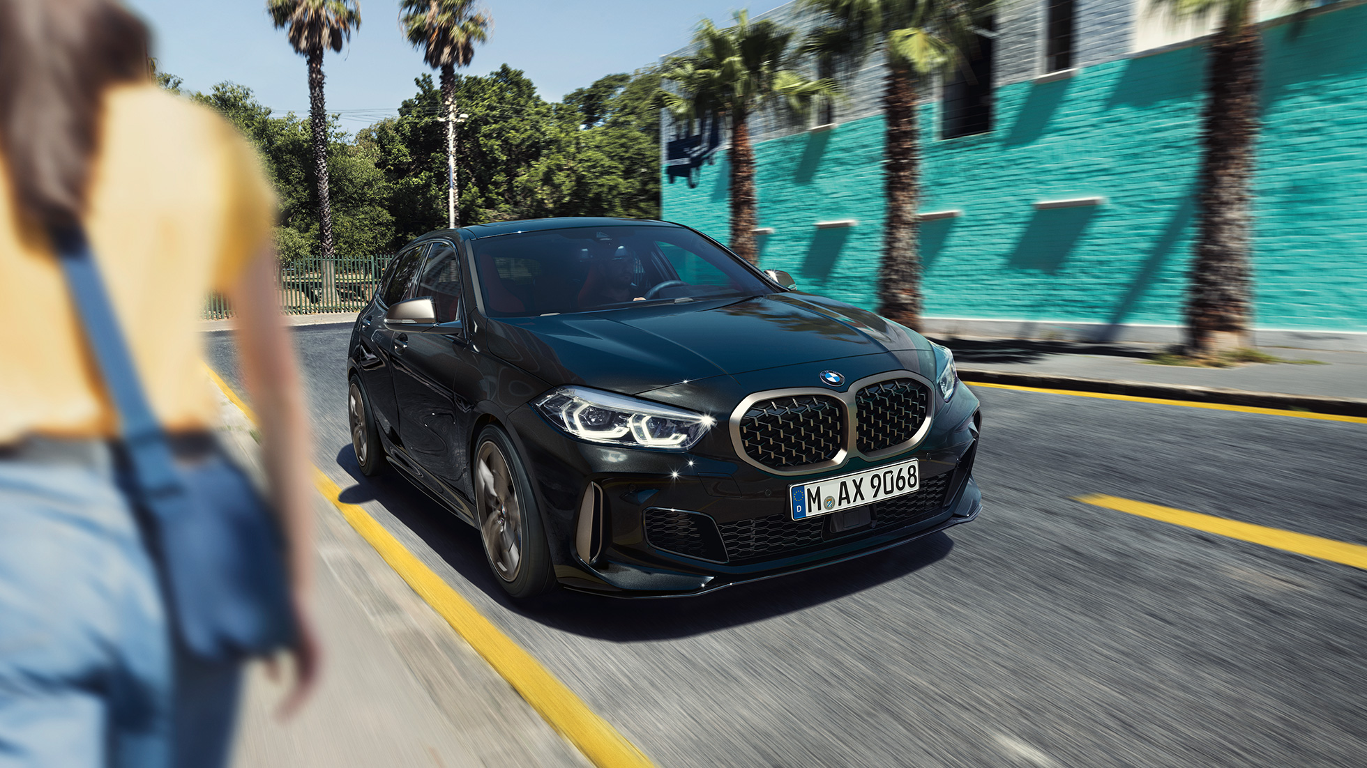 BMW M135i xDrive, vista anteriore a tre quarti F40. BMW M135i xDrive in Sapphire Black metallizzato, vista anteriore a tre quarti con donna in primo piano F40.
