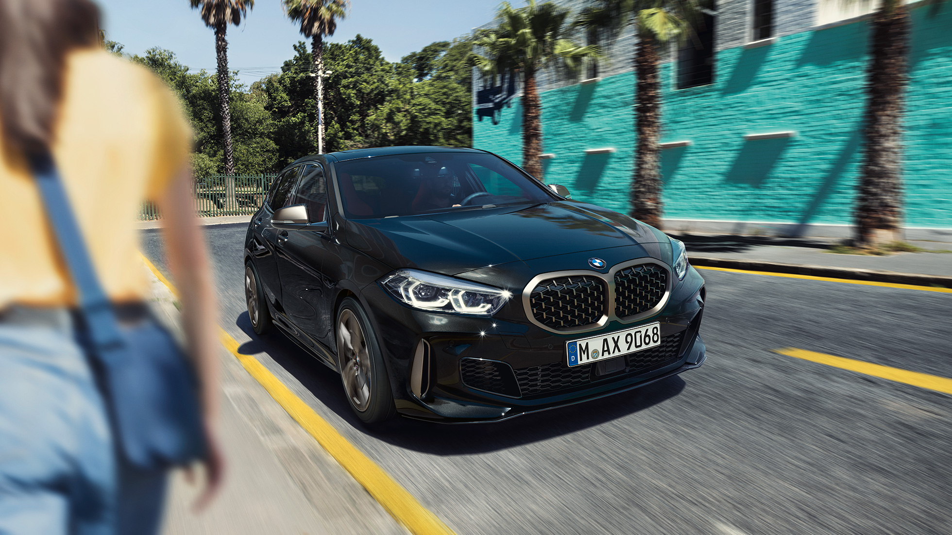 BMW M135i xDrive, vista anteriore a tre quarti F40. BMW M135i xDrive in Sapphire Black metallizzato, vista anteriore a tre quarti con donna in primo piano F40.
