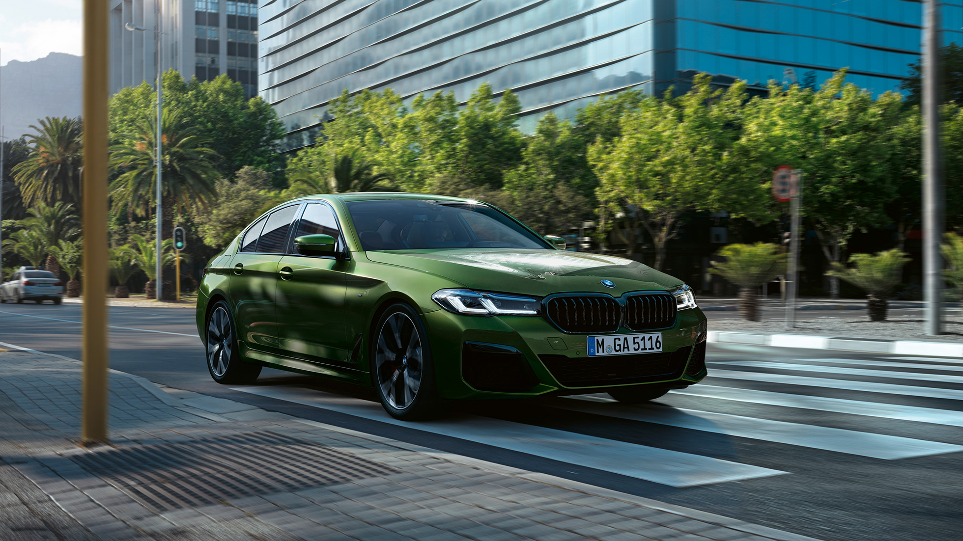 BMW M550i xDrive Berlina G30 LCI Facelift 2020 vista anteriore a tre quarti in movimento BMW M550i xDrive Berlina G30 LCI Facelift 2020 Hermes Green BMW Individual metallizzato vista anteriore a tre quarti in movimento su sfondo urbano