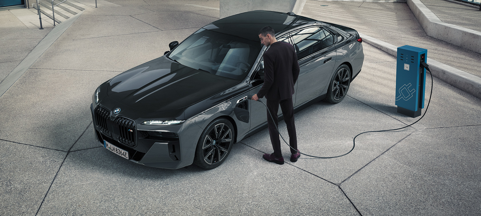 BMW M760e xDrive Berlina G70 Pacchetto M Performance Vista anteriore a tre quarti con uomo con cavo di ricarica in mano vicino a una stazione di ricarica e BMW M760e xDrive Berlina G70 con pacchetto M Performance BMW vernice BMW Individual bicolore in Sapphire Black metallizzato e BMW Individual Dravit Grey metallizzato