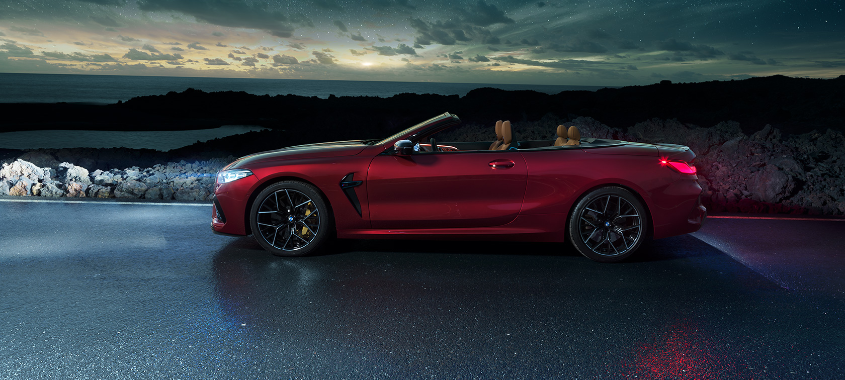 BMW M8 Competition Cabrio, vista laterale F91. BMW M8 Competition Cabrio in Motegi Red metallizzato con capote aperta, vista laterale, in sosta su una strada costiera sotto il cielo stellato F91.