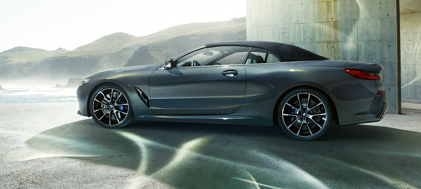 BMW M850i xDrive Cabrio, vista laterale G14. BMW M850i xDrive Cabrio in Dravit Grey metallizzato con capote chiusa, vista laterale davanti a catena di colline G14.