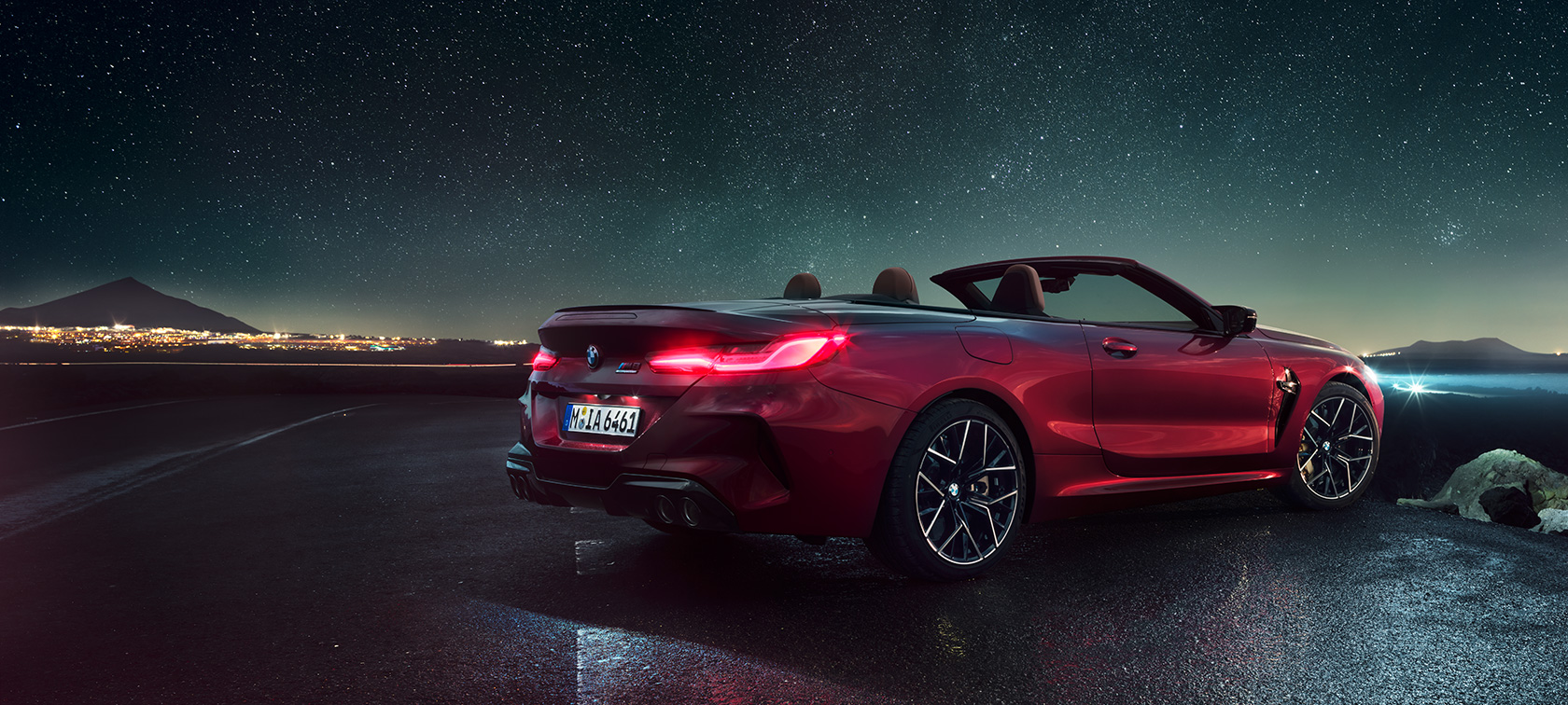 BMW M8 M Competition Cabrio, vista posteriore di tre quarti F91. BMW M8 Competition Cabrio in Motegi Red metallizzato con capote aperta, vista posteriore di tre quarti, in sosta sotto il cielo stellato F91.