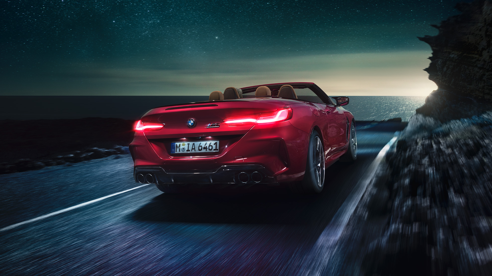 BMW M8 M Competition Cabrio, vista posteriore di tre quarti F91. BMW M8 Competition Cabrio in Motegi Red metallizzato con capote aperta, vista posteriore di tre quarti, in marcia di notte su strada costiera F91.