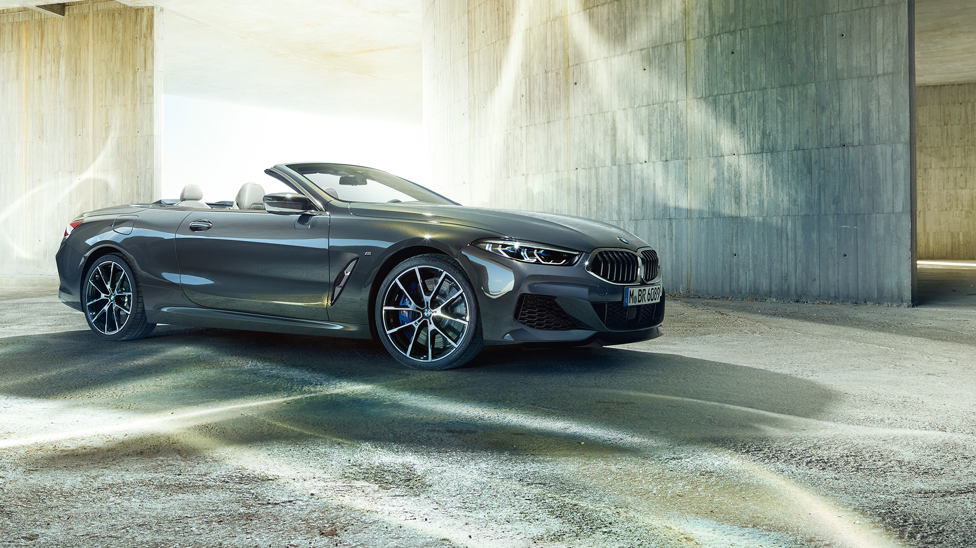 BMW M850i xDrive Cabrio, vista laterale di tre quarti G14. BMW M850i xDrive Cabrio in Dravit Grey metallizzato con capote aperta, vista laterale di tre quarti, in sosta davanti a un edificio in calcestruzzo G14.