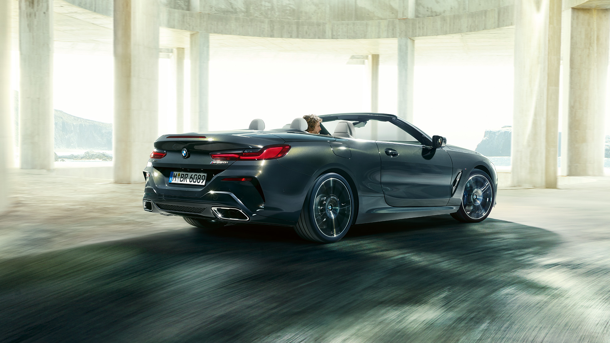 BMW M850i xDrive Cabrio, vista posteriore di tre quarti G14. BMW M850i xDrive Cabrio in Dravit Grey metallizzato con capote aperta, vista posteriore di tre quarti, in marcia attraverso un edificio in calcestruzzo G14.