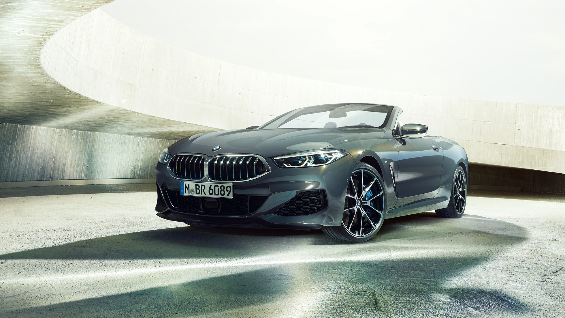 BMW M850i xDrive Cabrio, vista frontale di tre quarti G14. BMW M850i xDrive Cabrio in Dravit Grey metallizzato con capote aperta, vista frontale di tre quarti, in sosta in un edificio in calcestruzzo G14.