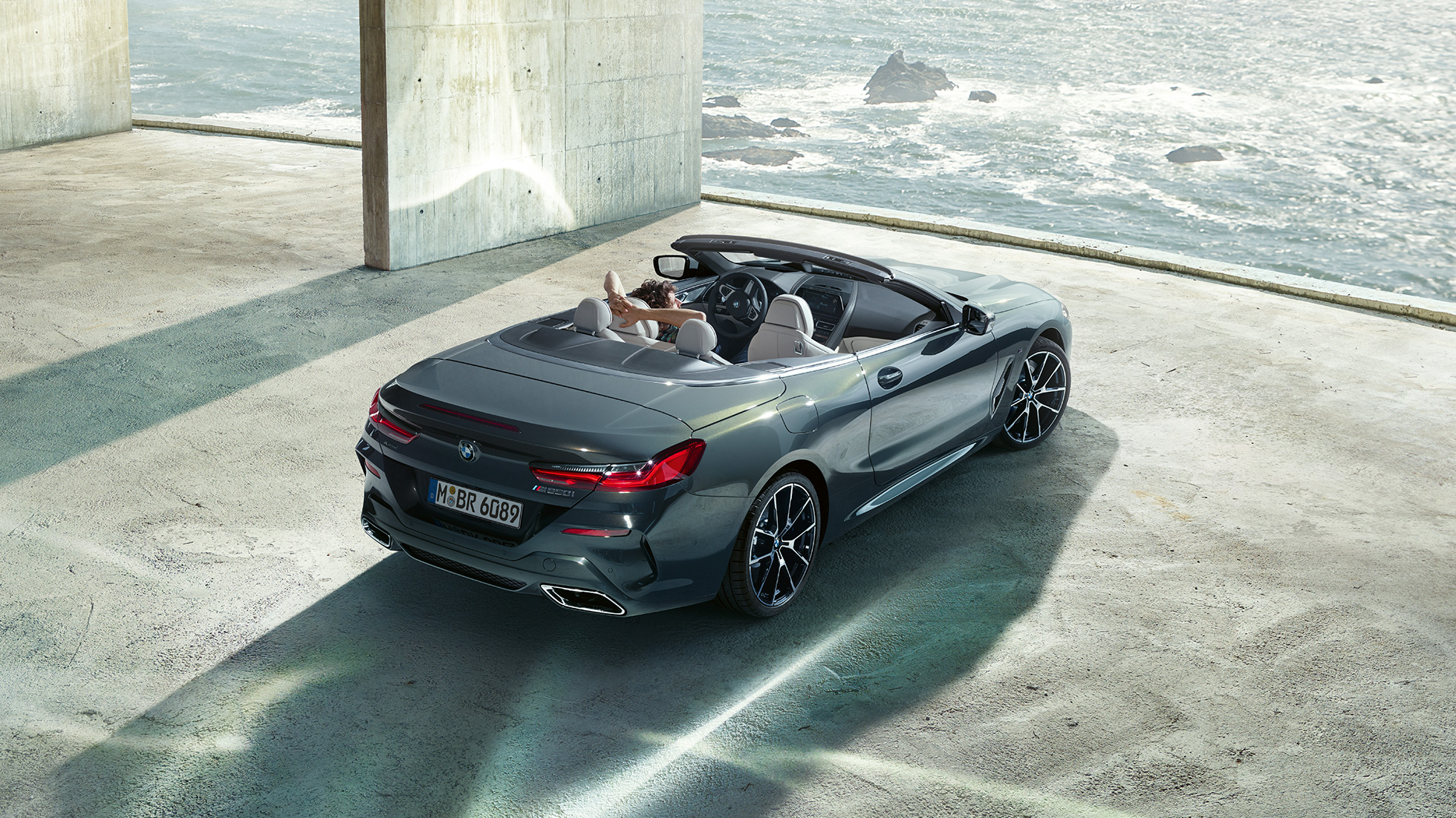 BMW M850i xDrive Cabrio, prospettiva a volo d’uccello G14. BMW M850i xDrive Cabrio in Dravit Grey metallizzato con capote aperta, prospettiva a volo d’uccello, parcheggiata in edificio di calcestruzzo davanti all’oceano G14.