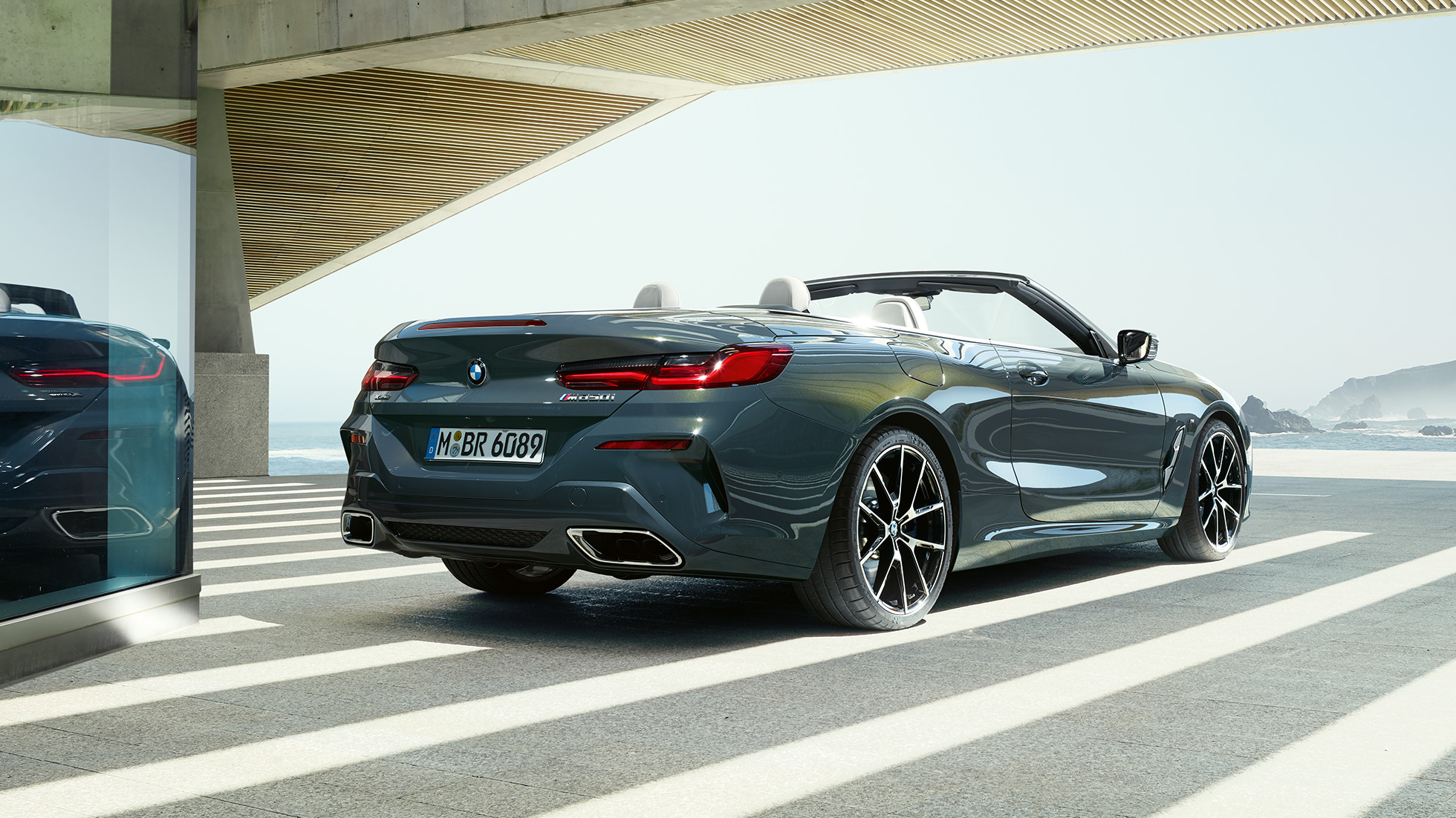 BMW M850i xDrive Cabrio, vista posteriore di tre quarti G14. BMW M850i xDrive Cabrio in Dravit Grey metallizzato con capote aperta, vista posteriore di tre quarti, parcheggiata con sguardo sull’oceano G14.