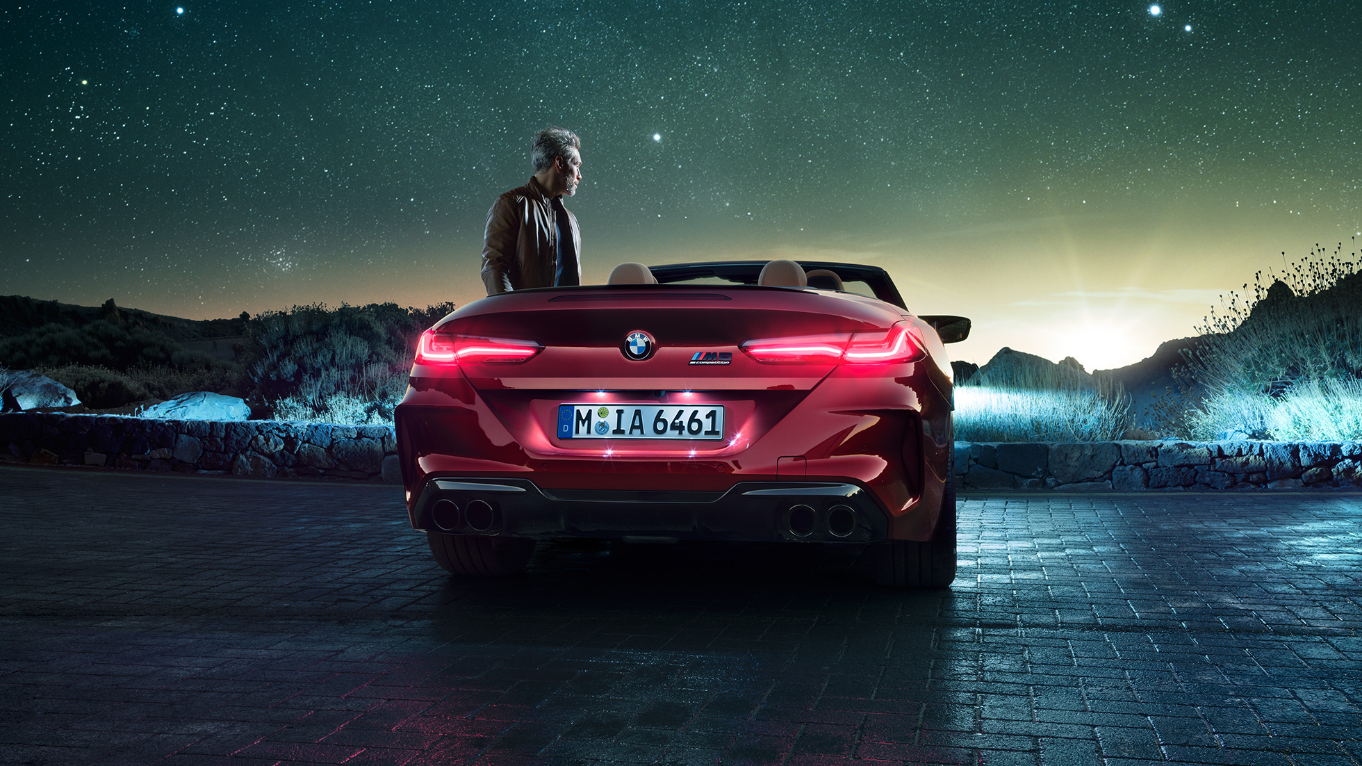 BMW M8 Competition Cabrio, vista posteriore F91. BMW M8 Competition Cabrio in Motegi Red metallizzato con capote aperta, vista posteriore, in sosta con uomo sotto il cielo stellato F91.