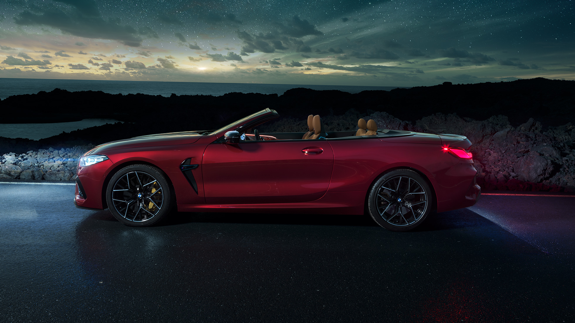 BMW M8 Competition Cabrio, vista laterale F91. BMW M8 Competition Cabrio in Motegi Red metallizzato con capote aperta, vista laterale, in sosta di notte sotto il cielo stellato F91.
