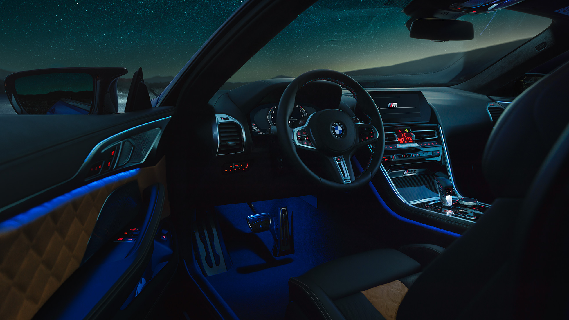Interni BMW M8 Competition Coupé F92. BMW M8 Competition Coupé, interni di notte con volante in pelle M e luce ambiente blu con porta del guidatore aperta F92.