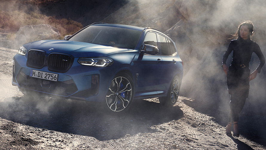 BMW X3 M Competition F97 LCI 2021 Vista anteriore a tre quarti ferma su sottofondo di sassi BMW X3 M Competition F97 LCI Facelift 2021 Frozen Marina Bay Blue metallizzato Vista anteriore a tre quarti ferma in salita in collina con modella