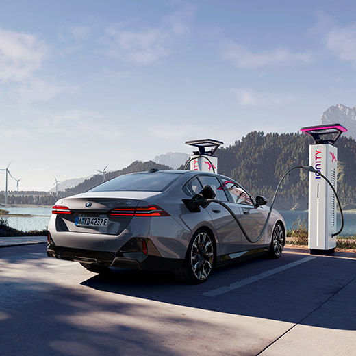 BMW i5 eDrive40 G60 vicina a una colonnina di ricarica BMW i5 eDrive40 G60 vicina a una colonnina di ricarica con un lago sullo sfondo.
