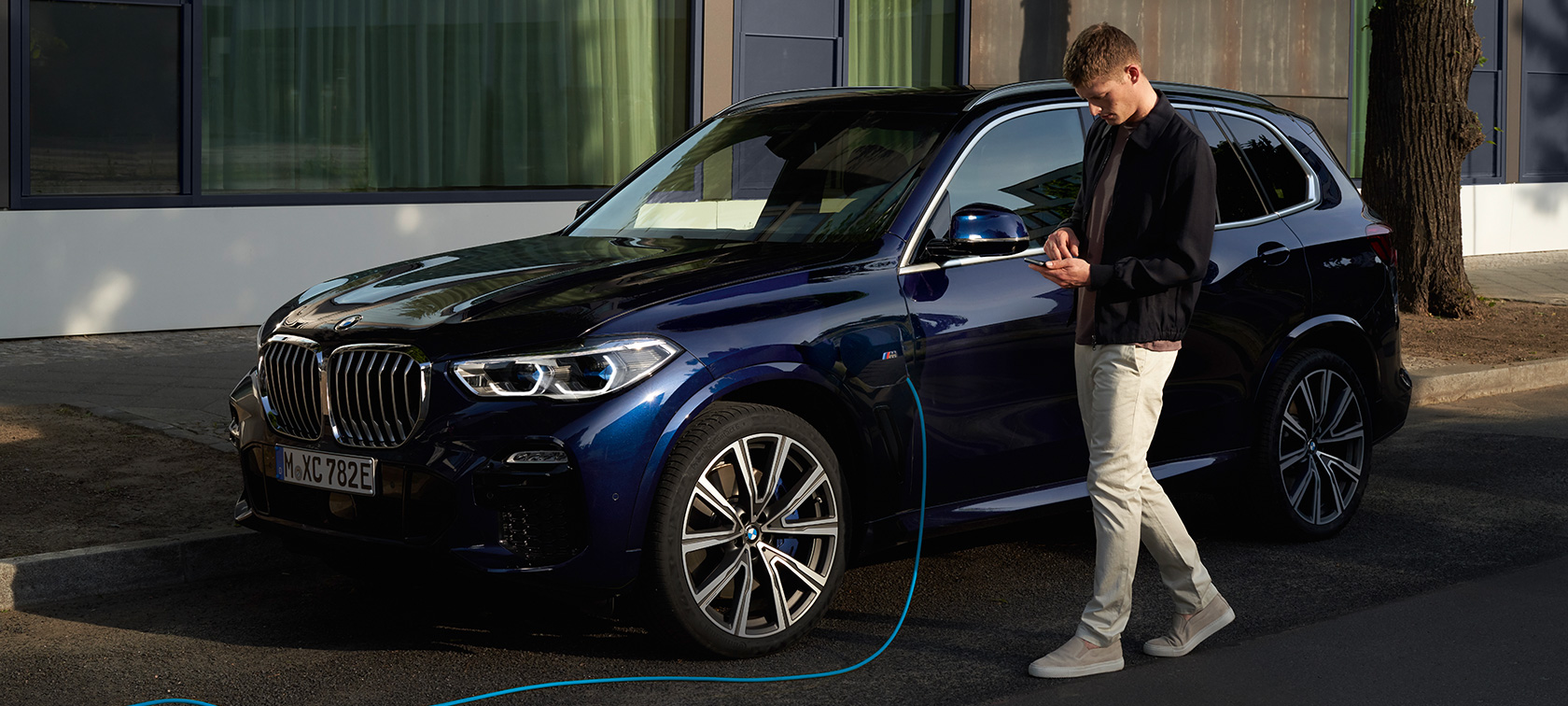 Un uomo utilizza l’app My BMW sul proprio smartphone Un uomo utilizza l’app My BMW di BMW ConnectedDrive sul proprio smartphone mentre la sua BMW X5 ibrida Plug-in si ricarica