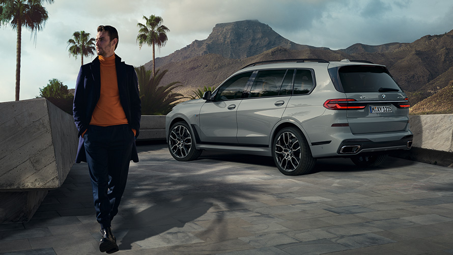 Un uomo davanti a una BMW X7 (G07) parcheggiata Remote Control Parking Remote Control Parking Un uomo davanti a una BMW X7 (G07) in vista posteriore a tre quarti parcheggiata con palme e sfondo di colline
