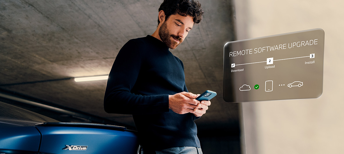 Un uomo è appoggiato al posteriore di una BMW con lo sguardo sul suo smartphone Remote Software Upgrade Un uomo è appoggiato al posteriore di una BMW con lo sguardo sul suo smartphone