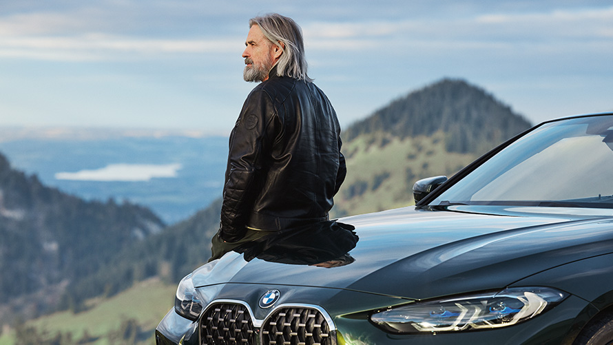 Un uomo appoggiato ad una BMW con sfondo collinare Accessori BMW primavera ed estate Un uomo appoggiato ad una BMW con sfondo collinare
