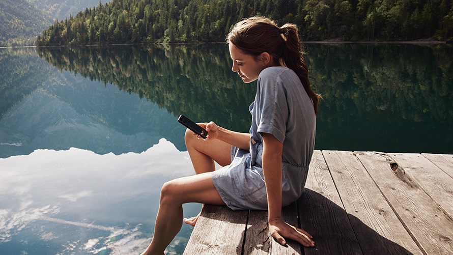 Una donna seduta su un pontile guarda il proprio smartphone Accessori BMW primavera ed estate Una donna seduta su un pontile guarda il proprio smartphone