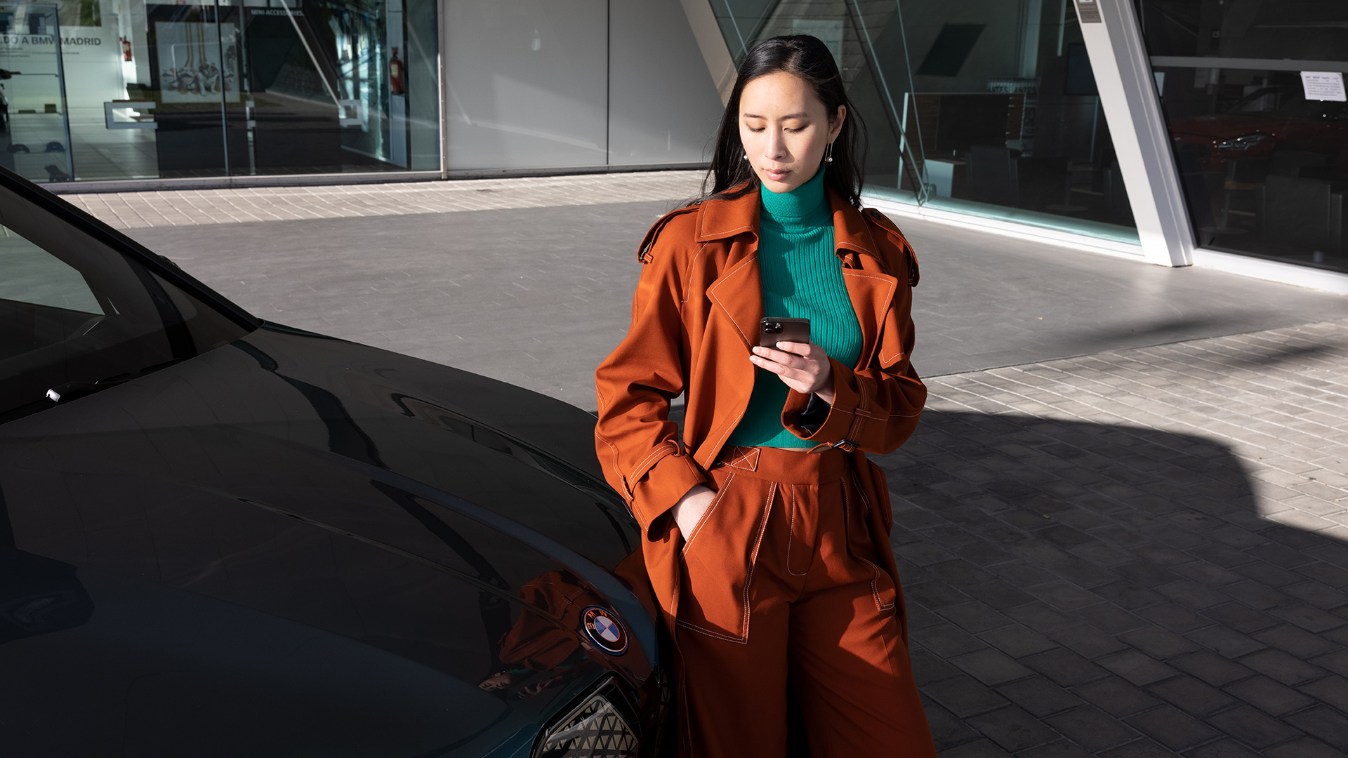 Donna in abito arancione si trova accanto a una BMW di fronte a un edificio e guarda il suo smartphone